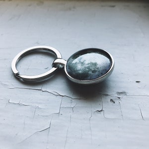 May include: A silver key chain with a round, glass charm featuring a black and white image of the moon.
