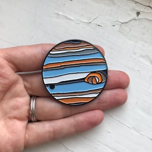 May include: A round, blue, orange, and white enamel pin with a black outline. The pin depicts a stylized image of Jupiter, with its iconic red spot and swirling bands of clouds.