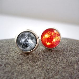 May include: A pair of silver stud earrings with a black and white moon image on one earring and a red and orange sun image on the other earring.