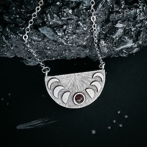 May include: A silver necklace with a crescent moon pendant featuring a red gemstone in the center. The pendant is surrounded by smaller crescent moons.