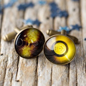 May include: A pair of cufflinks with a vintage design. The cufflinks are made of metal and have a round, glass-like surface with a yellow and brown pattern.