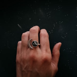 May include: A silver ring with a crescent moon design and a black stone in the center. The ring is on a person's finger.