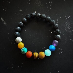 May include: A black lava rock bracelet with colorful beads representing the planets in our solar system. The beads are in order from the sun outward: yellow, orange, red, brown, yellow, blue, blue, purple.