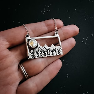 May include: Silver-toned mountain range pendant necklace. The rectangular pendant features a detailed mountain scene with a moon and trees. The necklace is held by a silver chain. The pendant is approximately 1.5 inches wide.