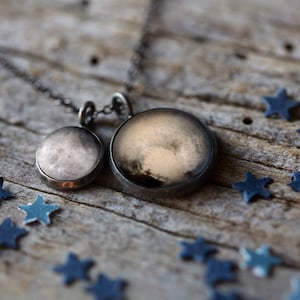 May include: A silver necklace with two round pendants featuring images of the moon. The larger pendant is on a black chain and the smaller pendant is on a silver chain. The pendants are on a wooden surface with blue stars scattered around them.