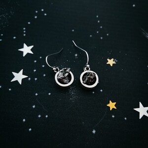 May include: A pair of silver earrings with a circular design. Each earring features a dark gray stone encased in a silver frame.