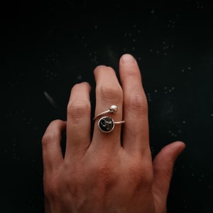 May include: A silver ring with a black stone set in a circular bezel. The ring is designed to wrap around the finger and has a small silver ball at the end of the wrap.