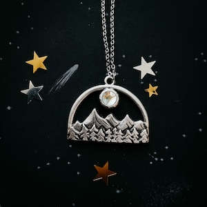 May include: A silver necklace with a mountain range pendant and a clear crystal stone. The pendant is shaped like a half circle and features a detailed mountain range design. The necklace is on a black background with gold stars.