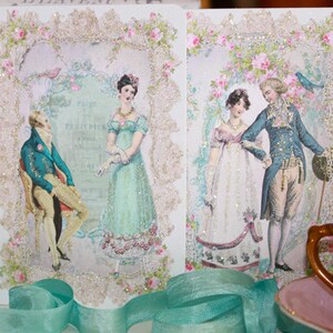 May include: Two decorative cards featuring vintage illustrations of couples in period clothing. The cards are framed with glittery lace and floral accents. The illustrations depict men in blue coats and women in pastel gowns, evoking a romantic, historical theme.