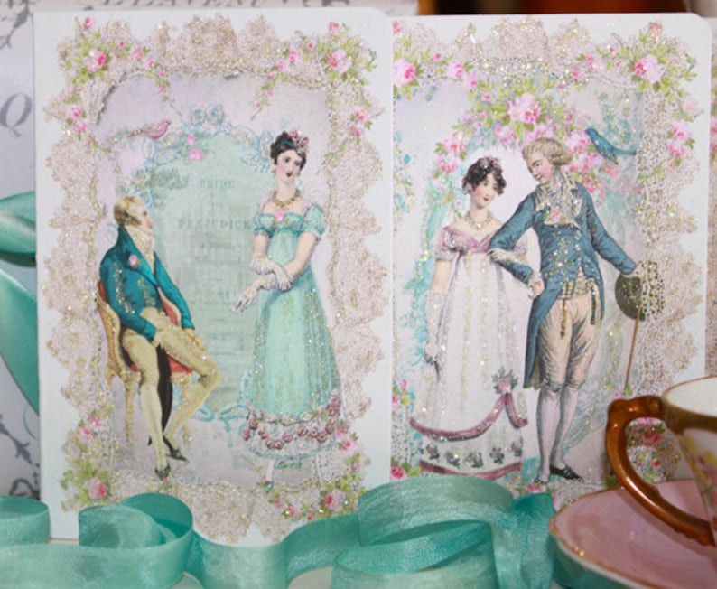 May include: Two decorative cards featuring vintage illustrations of couples in period clothing. The cards are framed with glittery lace and floral accents. The illustrations depict men in blue coats and women in pastel gowns, evoking a romantic, historical theme.