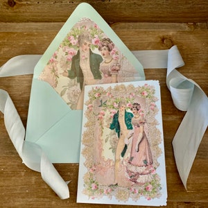 May include: A light blue envelope with a floral interior and a matching card featuring a vintage illustration of a couple in period clothing. A white ribbon is draped across a wooden surface.