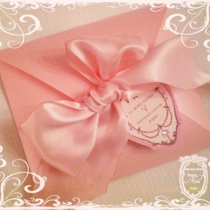 May include: A pink satin ribbon tied in a bow on a pink envelope with a white heart-shaped tag that says "La Noci Papille", "Paris Boutique Couture", and "Paris".