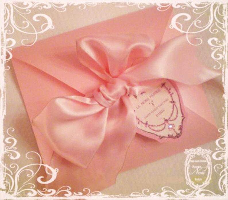 May include: A pink satin ribbon tied in a bow on a pink envelope with a white heart-shaped tag that says "La Noci Papille", "Paris Boutique Couture", and "Paris".