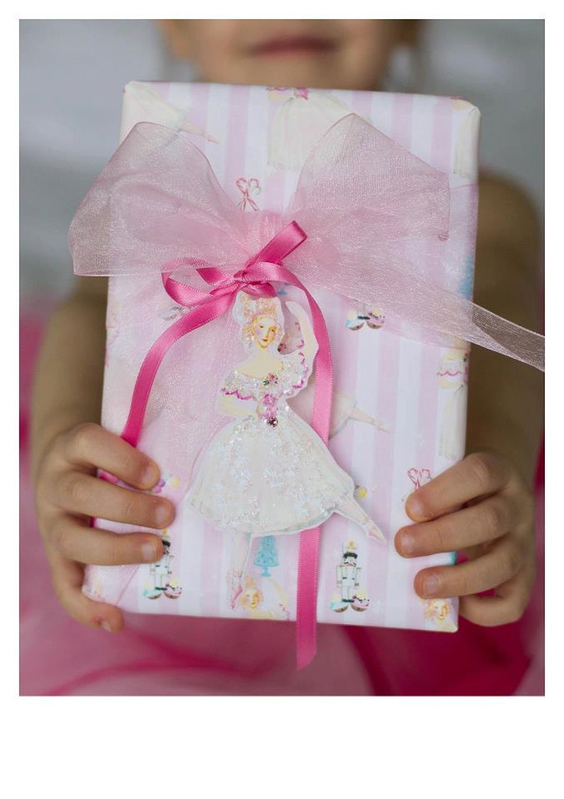 May include: A pink and white striped gift box with a pink tulle bow and a ballerina cutout. The ballerina is wearing a white dress with glitter and a pink ribbon.