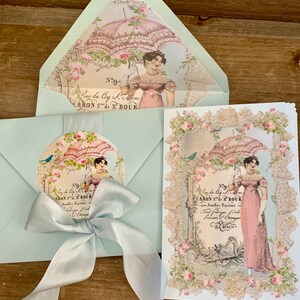 May include: A set of vintage-style stationery featuring a light blue envelope with a matching bow, a card, and a circular sticker. The designs include a woman with a parasol, floral accents, and French text.