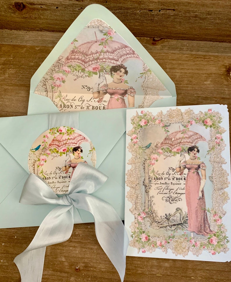 May include: A set of vintage-style stationery featuring a light blue envelope with a matching bow, a card, and a circular sticker. The designs include a woman with a parasol, floral accents, and French text.