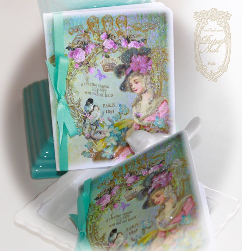 May include: Two vintage-style decorative boxes with a teal ribbon and a floral design. The boxes feature a woman in a hat and a bird with the text "a chaque oiseau son nid est beau PARIS 1897".