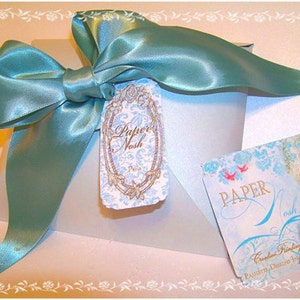 May include: A light blue satin ribbon tied in a bow on top of a white box with a decorative paper tag that says "Papier Nash" and a smaller tag that says "PAPER Nash Creative Printing & Artful Details, Stationery Designs from Paris".