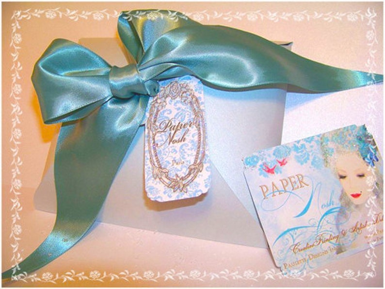 May include: A light blue satin ribbon tied in a bow on top of a white box with a decorative paper tag that says "Papier Nash" and a smaller tag that says "PAPER Nash Creative Printing & Artful Details, Stationery Designs from Paris".