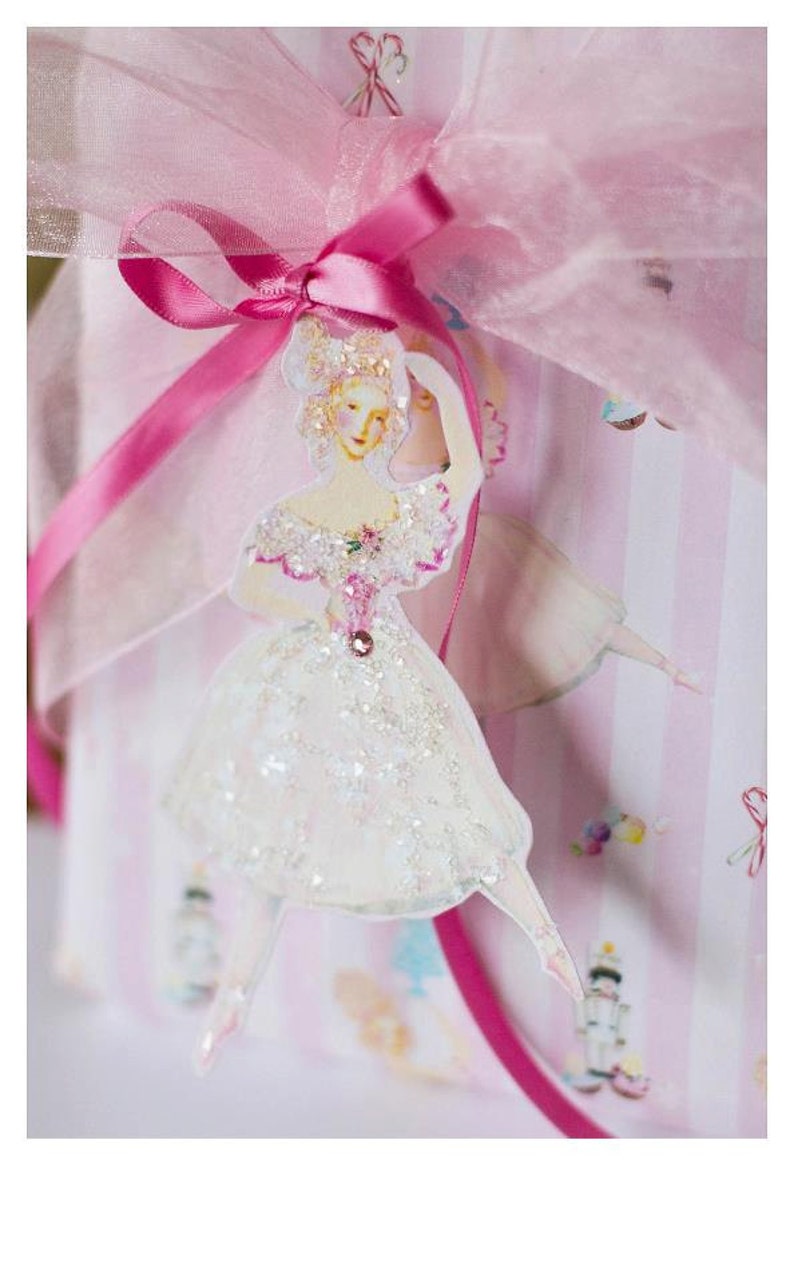 May include: A pink tulle bow with a pink ribbon tied around it. A white ballerina figurine with a sparkly white dress is attached to the bow.