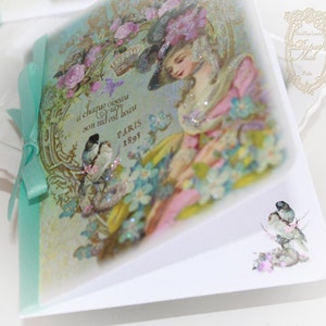 May include: A vintage-style card with a light blue background featuring a woman in a pink dress and a hat. The card has a gold border and text that reads "A chaque oiseau son nid est beau Paris 1895".