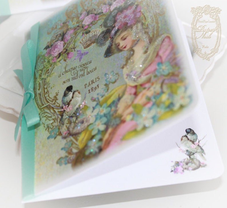 May include: A vintage-style card with a light blue background featuring a woman in a pink dress and a hat. The card has a gold border and text that reads "A chaque oiseau son nid est beau Paris 1895".
