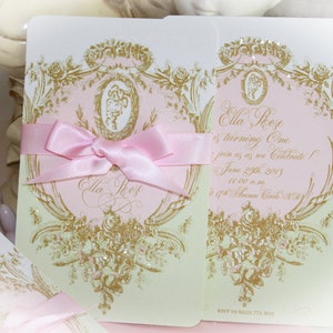Puede incluir: Una invitación rosa y verde con detalles dorados y una cinta de satén rosa. La invitación dice "Ella Rose cumple un año, ¡únase a nosotros para celebrar! 29 de junio de 2013, de 11:00 a.m. a 1:00 p.m.  RSVP a Kelly 773-3013"