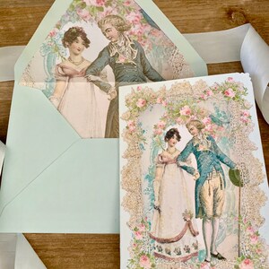 May include: A light blue envelope and card set featuring a vintage illustration of a couple in period clothing. The card has a decorative floral border with gold glitter accents. A white ribbon is draped across the wooden surface.