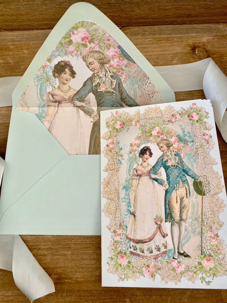 May include: A light blue envelope and card set featuring a vintage illustration of a couple in period clothing. The card has a decorative floral border with gold glitter accents. A white ribbon is draped across the wooden surface.
