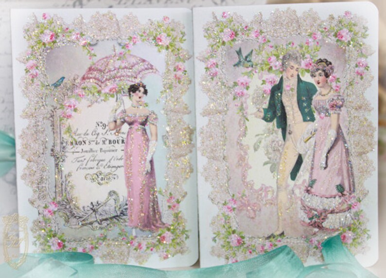 May include: A decorative journal featuring vintage illustrations of people in period clothing. The pages are framed with lace and floral accents, with glitter embellishments. The journal has a romantic, antique aesthetic.