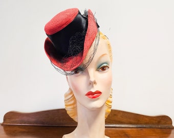 May include: A red and black hat with a black veil. The hat is sitting on a mannequin head.