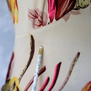 May include: Close-up of a cream-colored garment with a floral pattern in shades of pink, red, yellow, and brown. A pencil with a floral design is in the foreground. The garment appears to be a top or dress.