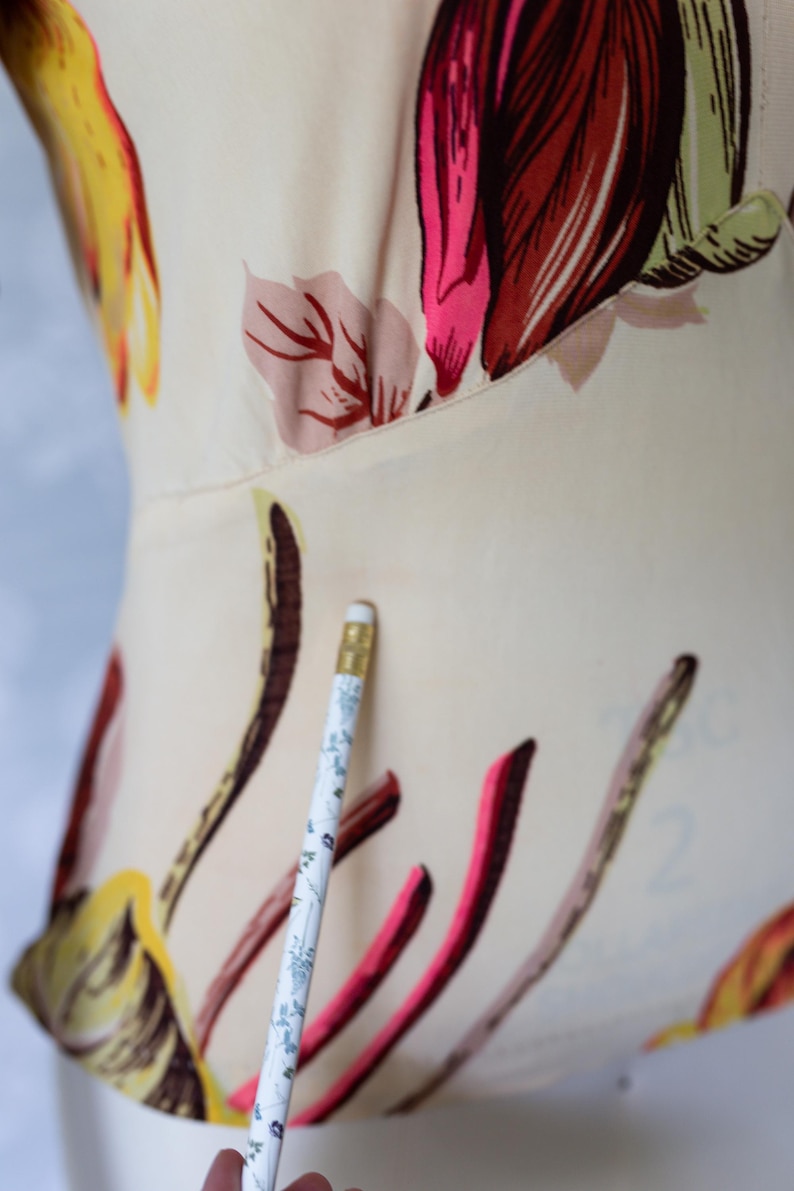 May include: Close-up of a cream-colored garment with a floral pattern in shades of pink, red, yellow, and brown. A pencil with a floral design is in the foreground. The garment appears to be a top or dress.