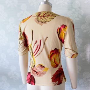 May include: A cream-colored blouse with a floral pattern featuring red, yellow, and burgundy tulip-like flowers and leaves. The short-sleeved top is displayed on a mannequin, showcasing the back view of the garment. The fabric appears to be lightweight.