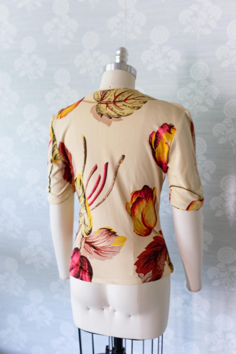 May include: A cream-colored blouse with a floral pattern featuring red, yellow, and burgundy tulip-like flowers and leaves. The short-sleeved top is displayed on a mannequin, showcasing the back view of the garment. The fabric appears to be lightweight.
