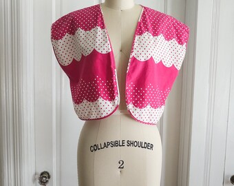 May include: A pink and white polka dot sleeveless cropped jacket with a scalloped hem. The jacket is on a dress form with the words "Collapsible Shoulder 2" printed on the front.