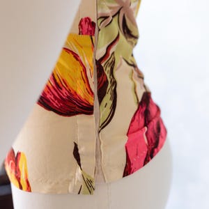May include: Close-up of a floral print garment, featuring a vibrant pattern of yellow, red, and pink flowers on a cream background. The fabric is visible, with seams and stitching details. The garment is displayed on a white mannequin.