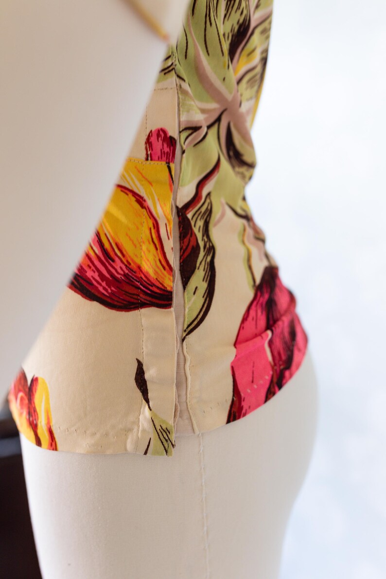 May include: Close-up of a floral print garment, featuring a vibrant pattern of yellow, red, and pink flowers on a cream background. The fabric is visible, with seams and stitching details. The garment is displayed on a white mannequin.