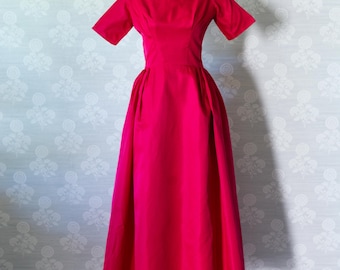 May include: A vibrant, fuchsia-colored vintage dress displayed on a mannequin. The dress features a boat neckline, short sleeves, and a full, flowing skirt. The dress is set against a patterned wallpaper background.