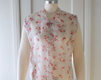May include: A vintage-style blouse with a sheer overlay, featuring a delicate floral pattern in shades of pink and red. The blouse has a bow at the neckline and is displayed on a mannequin. The sleeves are a solid cream color.