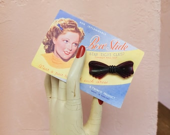 May include: A vintage advertisement for "Bow Slide" hair clips. The ad features a woman with a red bow in her hair and the text "NON INFLAMMABLE Bow Slide STAY TIGHT CLASP CANNOT FALL OUT Wear a pair to each dress A TWINCO PRODUCT MADE IN ENGLAND".