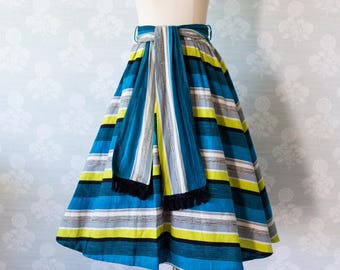 May include: A full-circle skirt with a blue, white, yellow, and black striped pattern. The skirt has a matching blue, white, and yellow striped sash tied around the waist.