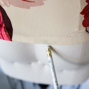 May include: Close-up of a floral print fabric on a dress form. The fabric features large pink, red, and yellow flowers on a cream background. A pencil with floral designs is positioned near the fabric.