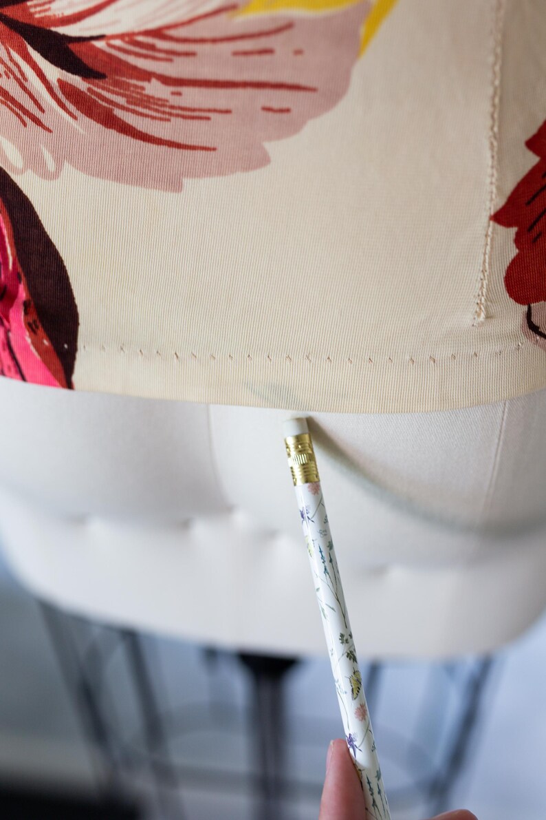 May include: Close-up of a floral print fabric on a dress form. The fabric features large pink, red, and yellow flowers on a cream background. A pencil with floral designs is positioned near the fabric.