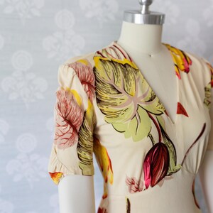 May include: Cream-colored dress with a V-neck and short sleeves. The dress features a vibrant floral pattern in shades of red, yellow, and green. The dress is displayed on a mannequin.