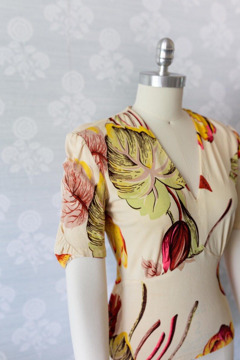 May include: Cream-colored dress with a V-neck and short sleeves. The dress features a vibrant floral pattern in shades of red, yellow, and green. The dress is displayed on a mannequin.