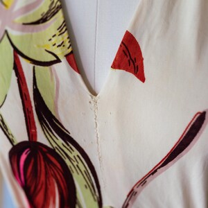May include: Close-up of a cream-colored dress with a V-neckline. The fabric is adorned with a floral pattern featuring red, pink, green, and brown elements. The dress is displayed on a mannequin.