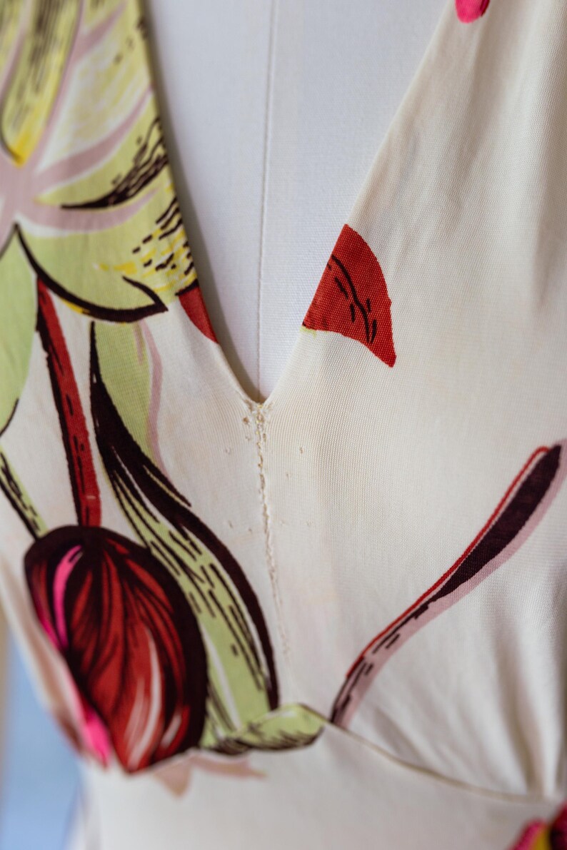 May include: Close-up of a cream-colored dress with a V-neckline. The fabric is adorned with a floral pattern featuring red, pink, green, and brown elements. The dress is displayed on a mannequin.