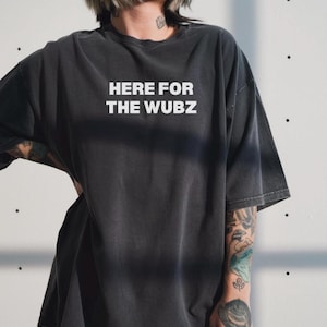 May include: Dark gray t-shirt with the text "HERE FOR THE WUBZ" in white block letters. The shirt has a slightly faded appearance and a relaxed fit. The model is wearing the shirt.