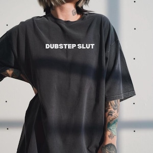 May include: Dark gray oversized t-shirt with the text "DUBSTEP SLUT" in white block letters. The shirt has a slightly faded, vintage look. The short-sleeved tee is a casual, unisex garment.